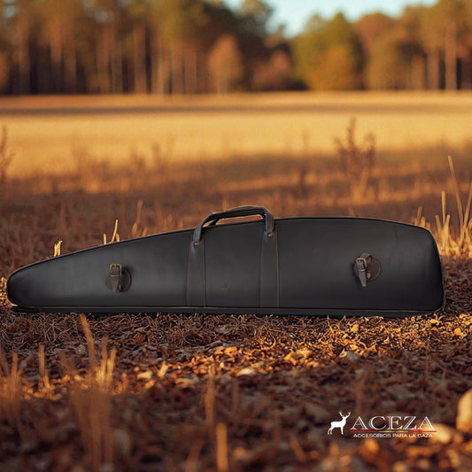 Funda de rifle Aceza en cuero marrón oscuro apoyada en un campo abierto con hierba seca y árboles al fondo.